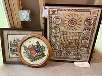 Three needlepoint framed artworks and needlepoint calendar displayed on a surface, including a large calendar piece with months and days, round artwork of children on fence, and house illustration framed piece.