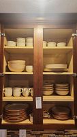 Upper cabinet with shelves holding stacks of Noritake stoneware: bowls, mugs, plates, ramekins, and serving dishes.