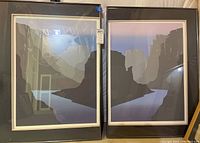 Pair of framed prints showing stylized landscape images with cliff and river scene under glass in black frames with white matte borders.