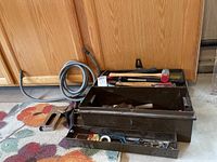 Photo showing open metal toolbox with various hand tools inside compartments and a coiled gray hose next to it on a flower patterned floor mat.