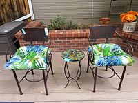 Two wrought iron armchairs with tropical patterned cushions and a small round side table with peacock design on patio deck.