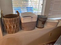 Four items displayed on window ledge: large woven basket with crisscross white string, two-tone round basket, metal pail with handles, and rectangular two-tone basket