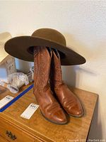Photo showing pair of brown Ariat leather boots with decorative stitching on shaft, pointed toes, and angled heels with brown felt wide-brim cowboy hat resting on boots