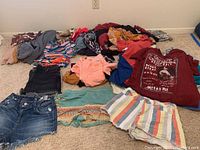 Wide shot showing various women's clothing items including shorts, tops, and dresses laid on the floor in assorted colors and styles.