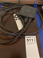 Apple TV device with remote control and HDMI cable, showing scratches and signs of wear, placed on dark surface with MaxSold lot tag visible.