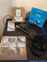 Photo showing black JBL wireless earbuds inside charging case, USB charger, sticker or patch with Pittsburgh Steelers logo, Brenn Hill autographed CD with cowboy image, and blue box labeled '4 piece master kit for Apple AirPods'.