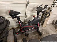 Side view of the exercise bike showing black frame with red and white accents, padded damaged seat, and pedals.
