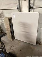 White Lifetime folding table, shown folded, leaning against a garage door with a concrete block beside it.