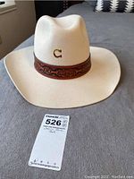 Front angle of white/cream wool western hat with brown leather band and gold 'C' logo on front.
