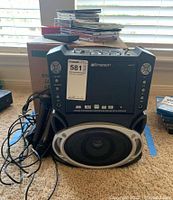 Front view of Emerson karaoke machine with control panel, built-in speaker, and wired microphone tangled on side. Stack of CDs/DVD cases and remote control on table behind.