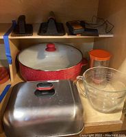 Photo of wooden cabinet shelf showing three meat presses, Thermpro meat thermometer, large red Cacinaware bowl with lid, glass measuring cup, orange-lidded container, and square grill lid.