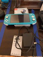 Front view of turquoise Switch Lite console with game case propped behind and AC adapter cable in front