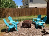 Four blue plastic Adirondack style outdoor chairs arranged around a firepit box on gravel in a backyard.
