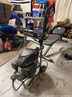 Photo showing Power Stroke gas pressure washer with handle, wheels, hose, and spray gun, surrounded by various other stored household items in cluttered background.