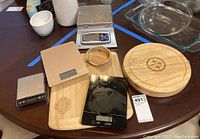 Overview of all items on round table showing four scales including a large metal one, three wooden items (two cutting boards and spoon rest), and glass cookware.