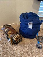 Blue sleeping bag rolled, secured with straps, with a white tag visible on the side. Pendleton wool blanket rolled and held with leather strap lying beside it.