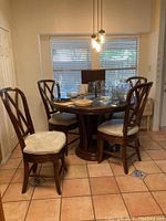 Full view of round wooden dining table with four matching chairs around it on tiled floor. Table contains various glassware and bowls. Light fixture overhead.