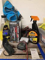 Photo showing NOCO Boost Plus jump starter in box, Armor All tire shine, jumper cables, Meguiar's wash & wax, blue microfiber cloth, flashlight, and a small black pouch on wooden surface.
