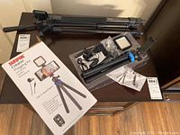 Photo of the lot showing the collapsed black tripod, Sunpak Creator Vlogging Kit box beside the kit components including a small LED light panel, microphone, various cables and mounts on a wooden surface.