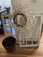 Two distressed white wood window frames with visible paint chipping leaning against a wall with a round flower wreath and wooden bucket positioned in front to show lot content.