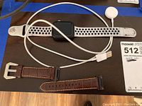 Apple Watch with Nike white and black perforated sport band, magnetic charging cable, and brown leather spare band laid out on dark surface.