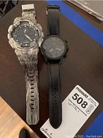 Both men's wristwatches side by side on a wooden surface, showing the full watches and bands. Fossil with black leather band, G-Shock with marbled gray resin band.