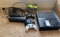 Photo showing Xbox One Model 1540 console, three Xbox wireless controllers, stack of Xbox game cases, and several cords on the floor.