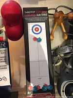 Photo showing tabletop curling game set in black box with two red and blue curling stones included, red ergonomic pillow support visible to the left, and wooden massage tool with knobs visible on right