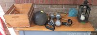 Wooden crate, weighted ball, dumbbells, lantern and carved wooden items shown on patio surface against brick wall.