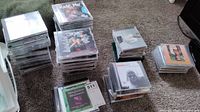 Multiple stacks of music CDs on carpet, over 90 total. Variety of jewel cases and album covers.