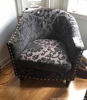 Front angled view of the gray floral damask upholstered accent chair showing the rounded back and nailhead trim along the arms and seat base.