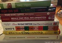 Stack of eight cookbooks showing titles related to vegan cooking, detox, Italian cuisine, and Anthony Bourdain.