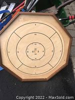 Top side of wooden octagonal game board showing Crokinole scoring rings and central hole, placed outdoors on pavement with garden tools in background.