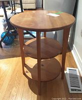 Light wood three tier round end table shown from angle highlighting circular tiers and slim legs on wood floor.