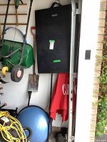 Showing black exercise mat hung vertically with green tape, blue balance ball on floor, garden tools and other items hanging nearby