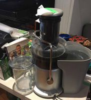 Silver and black juicer with clear juice container and grey pulp collector placed on a white table, next to a clear plastic juice jug and kitchen items in background.