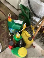 Photo showing green hand seeder, various bottles of fertilizers and weed killers, and part of a yellow container and handled gardening tools.