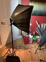 Side view of softbox light with white diffusion fabric, mounted on adjustable black tripod stand, showing electrical plug cable on floor.