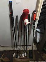 Eight pitch and putt golf clubs standing against a white wall with two headcovers on the clubs.