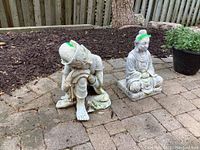 Two oriental resin garden figures side by side on outdoor brick surface with mulch bed background