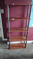 Front view of four-tier shelf unit showing medium brown laminate shelves with wear on edges and black/gray metal frame with simple square design side panels, positioned in a room with burgundy wall and carpeted floor.