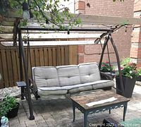 Full front view of metal patio swing with cushioned seating and wooden awning, placed on a stone patio with surrounding plants.