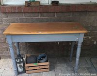 Front view of light wooden topped table with painted grey spindle legs and visible wear on top surface.