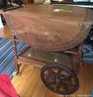 Side view showing drop leaf down and ornate carving detail on the folding leaf and turned legs. Also large wooden spoked front wheels and bottom shelf visible.