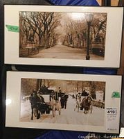 Two sepia prints in black plastic frames, visible measurements approx 10x20 inches, with old street scenes: one showing a pedestrian path with trees and a lamppost, the other snowy street with horse-drawn carriages.