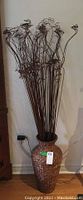Wicker vase with many long dried squiggly stalks next to a wooden cabinet against wall