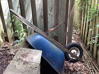 Side view of a blue metal wheelbarrow with wooden handles, resting against a wooden fence on dry leaves.