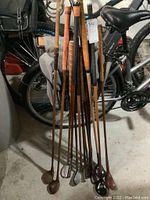 Full view of ten wooden golf clubs with hickory shafts, showing the clubs standing upright, handles wrapped with leather or natural grip, diverse head shapes with some heads wood and others metal.