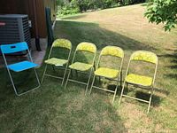 Five fold-up chairs positioned outside on grass: four green floral patterned chairs and one solid blue chair.
