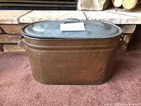 Front view of antique copper boiler with steel lid on carpeted floor in front of stone fireplace.